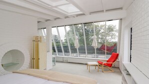 Un dormitorio con cama, una silla roja, una ventana con vistas a árboles y una pared de ladrillo blanco.