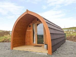 A small, wooden, semi-circular structure with a door and a window, a metal bucket, and a wooden bench outside.