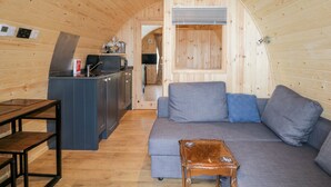 L’intérieur chaleureux d’un chalet avec un plafond en bois, un coin cuisine, un canapé gris et une petite table en bois.