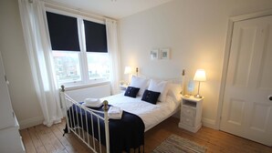 A bedroom with a bed, two bedside tables with lamps, a window with curtains, a radiator, and a door. - Quayside (Whitby)