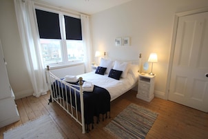 Un dormitorio con cama, dos mesitas de noche con lámparas, una ventana con cortinas, un radiador y una puerta.