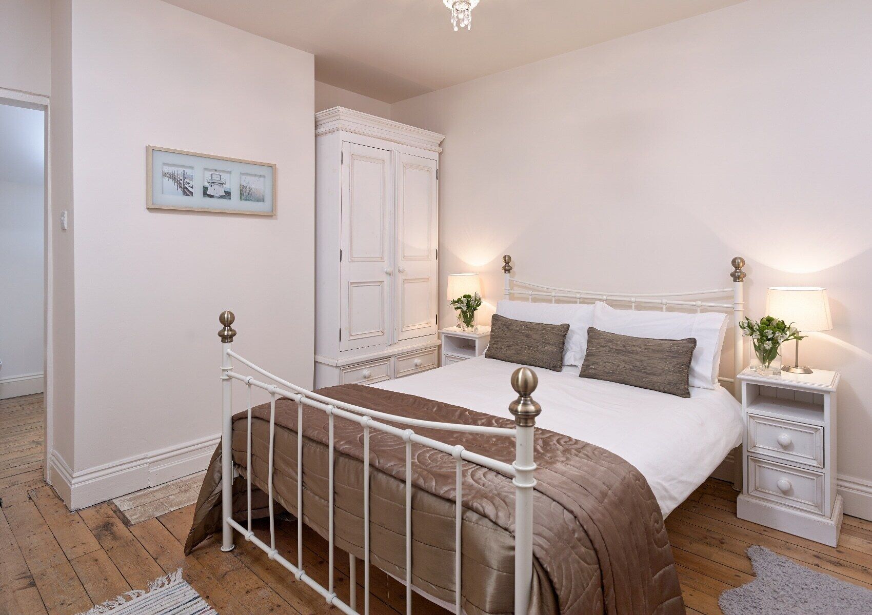 A bedroom with a white bed, a nightstand with a lamp, a wardrobe, and a framed picture on the wall.