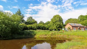A tranquil riverside scene with a small house featuring a sloped roof and a fenced garden. - The Stables (Frodsham)