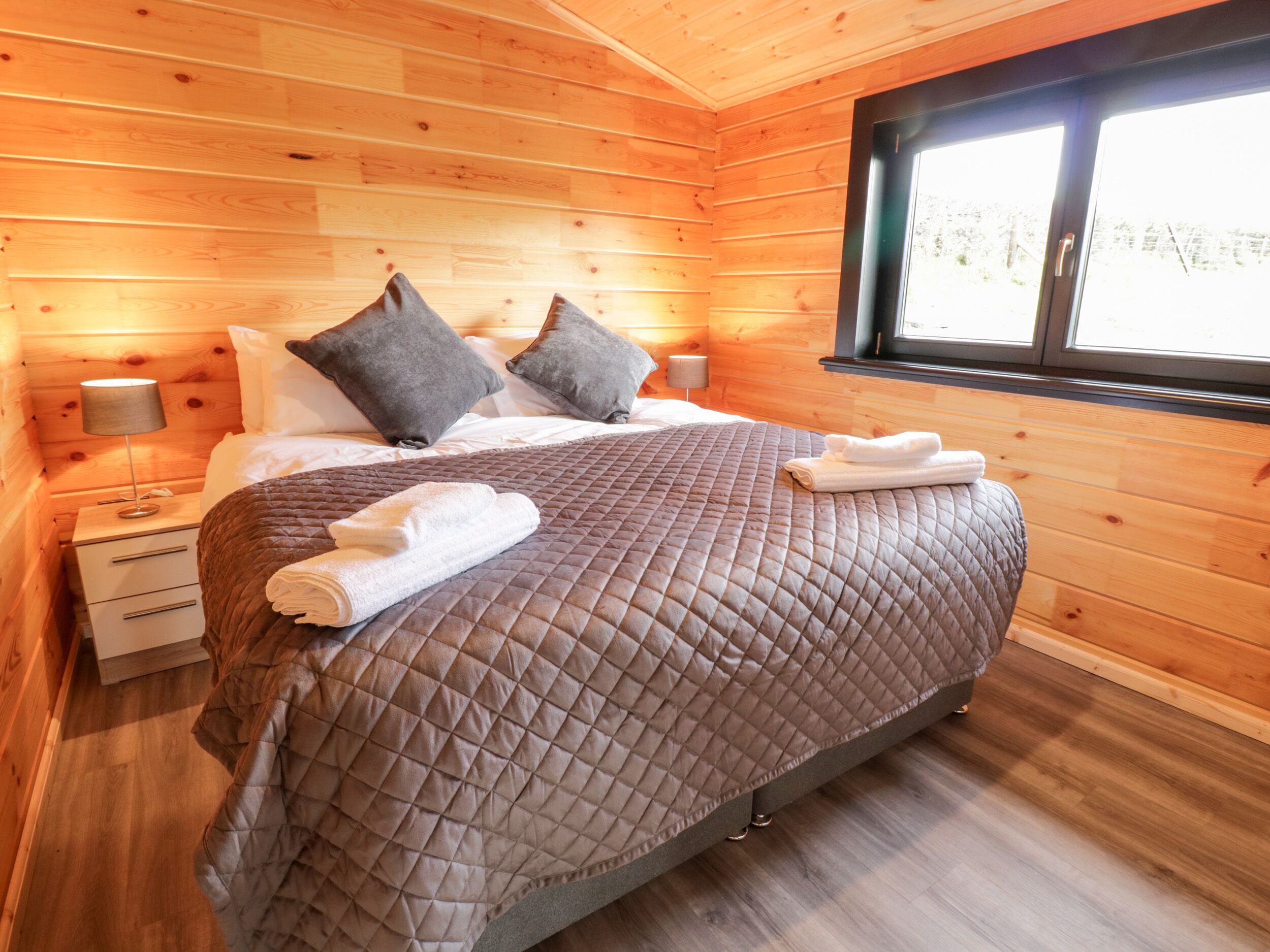 Ein Schlafzimmer mit Holzwänden, einem großen Bett, zwei Kissen, einem Nachttisch, einer Lampe und einem Fenster.