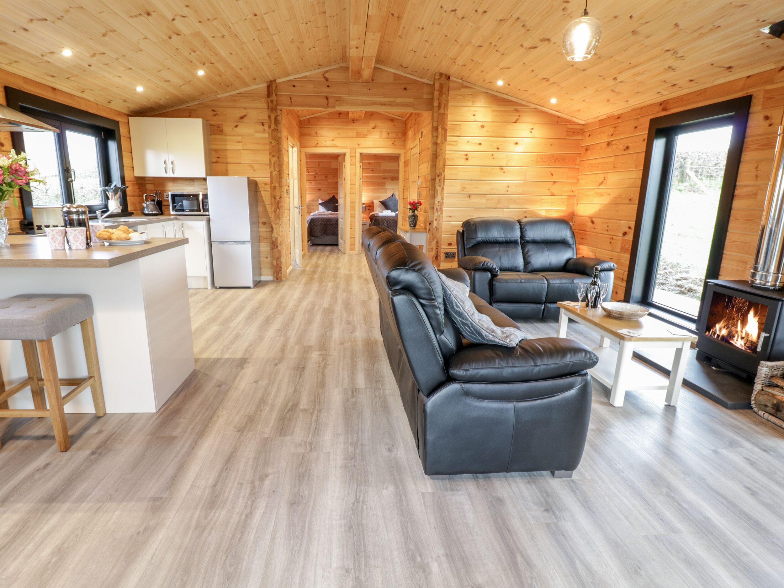 A log cabin interior with a kitchen, living area with leather sofas, a fireplace, and a large window.
