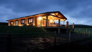 A wooden house with a covered porch and a fenced yard at dusk. - Bryn Eiddon Log Cabin (Machynlleth)