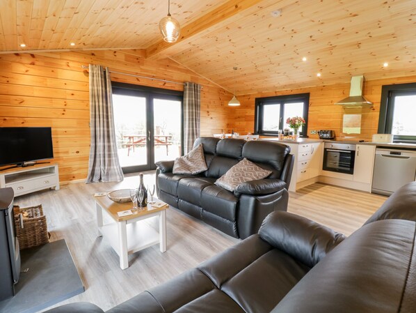 A cozy cabin interior with a black leather sofa, a TV, a dining table, and a kitchen area. - Bryn Eiddon Log Cabin (Machynlleth)