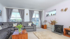 Ein modernes Wohnzimmer mit einem grauen Ecksofa, einem Holztisch, einem beigefarbenen Teppich, einem Kamin und Blick auf die Stadt durch große Fenster.