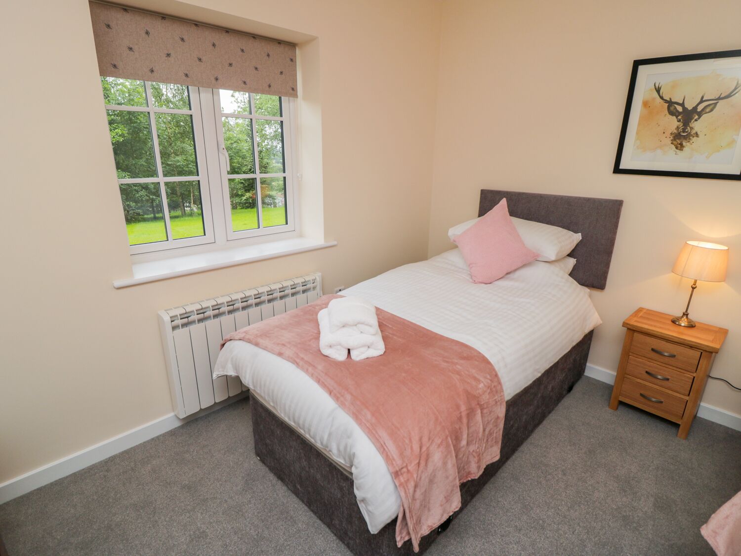 A bedroom with a bed, a bedside table with a lamp, a radiator, and a window with a view of greenery.