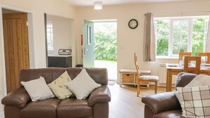 A living room with two brown leather sofas, a dining table with chairs, a clock on the wall, and a view of greenery outside. - Ryedale Country Lodges - Hazel Lodge (York)