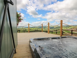 Un jacuzzi con terraza de madera y barandilla de cuerda, con vista a un paisaje pintoresco con colinas y cielo despejado.