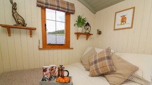 Ein gemütliches Schlafzimmer mit Bett, Nachttisch mit Kaffeemaschine und Croissants, Fenster mit Blick und Dekoration auf Regalen.