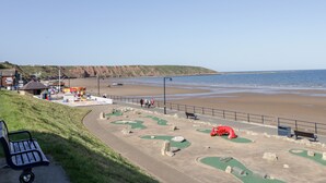 Eine Küstenlandschaft mit Sandstrand, einem kleinen Park mit Spielplätzen und einer Bank.