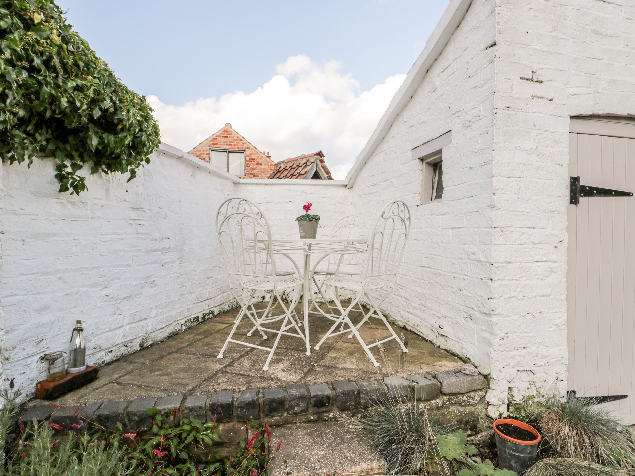 A white brick building with a small patio area featuring a metal table and chairs, surrounded by greenery and a potted plant.