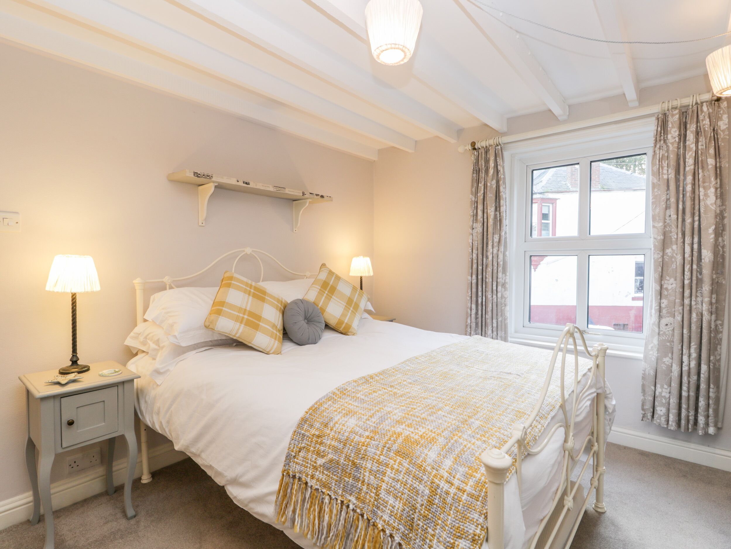 A neatly made bed with a white headboard, two bedside tables with lamps, a window with curtains, and a wall-mounted shelf above the bed.