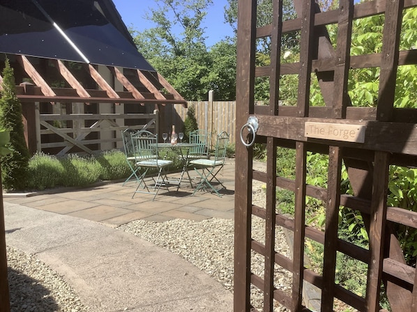 Una puerta de madera con un cartel que dice 'The Forge' da acceso a un patio con una mesa y sillas, rodeado de follaje.