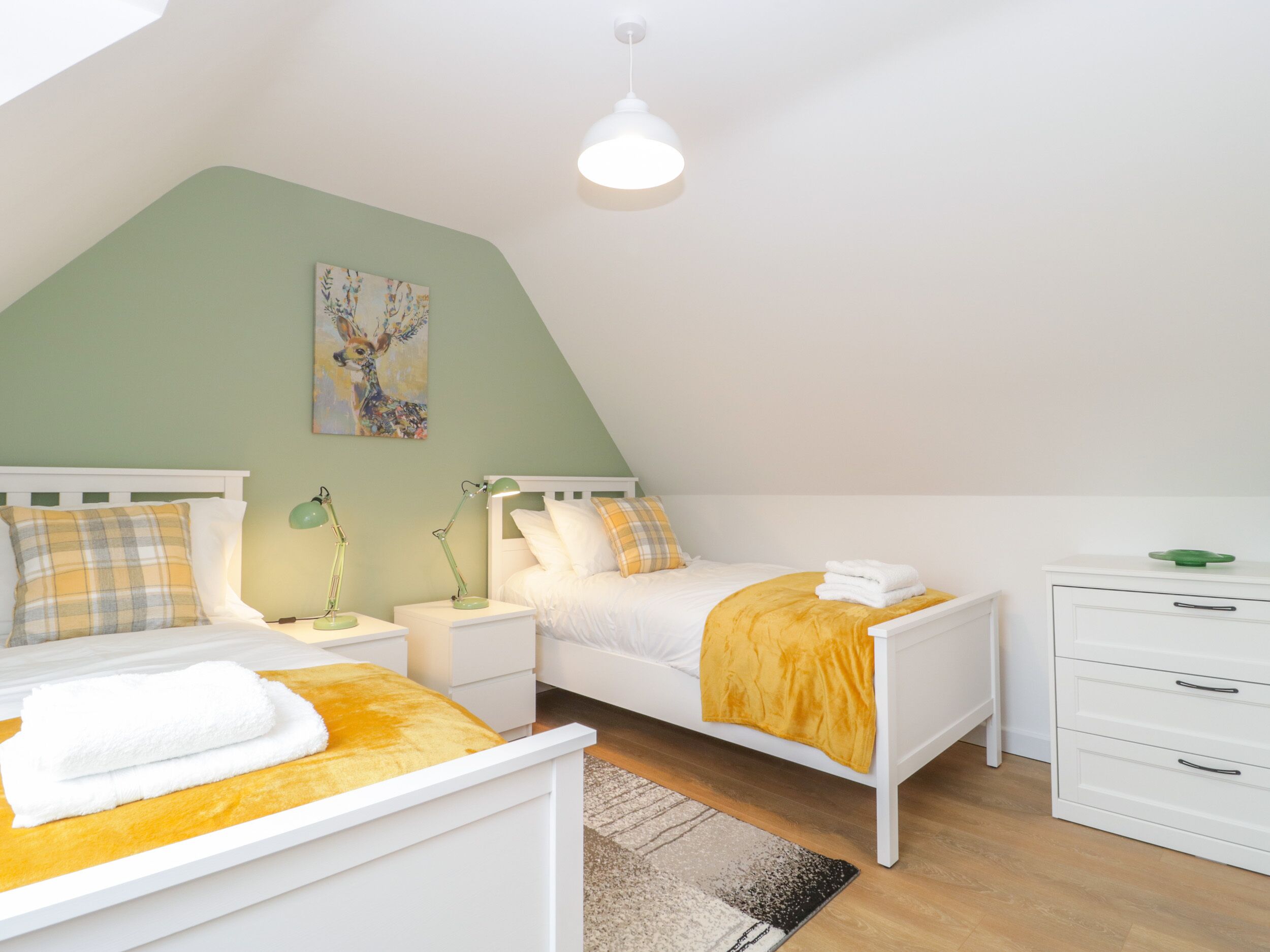 Et soveværelse med to senge, et natbord med lamper, en kommode og et billede på væggen.