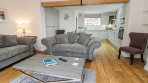 A living room with a grey sofa, a coffee table, and a red armchair. There is a kitchen in the background with white cabinets and a window. - Chalkstone Cottage (Bridlington)