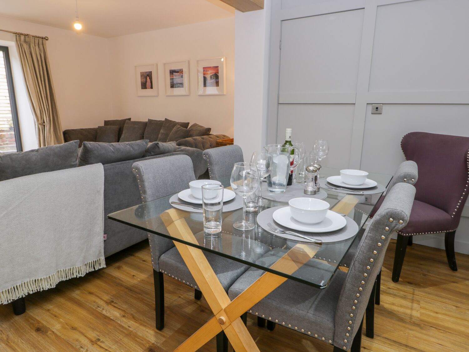 A modern living room with a glass dining table set for four, grey upholstered sofas, a purple armchair, and a white door in the background.