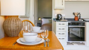 Una mesa de comedor dispuesta con platos, tazones y cubiertos blancos, una lámpara de mimbre y una cocina al fondo con armarios y electrodomésticos.