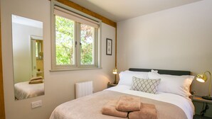 Un dormitorio con una cama grande, una ventana que da a árboles, una mesita de noche con una lámpara y un espejo en la pared.