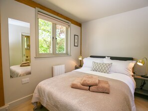 Un dormitorio con una cama grande, una ventana que da a árboles, una mesita de noche con una lámpara y un espejo en la pared.