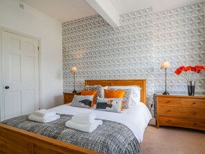A neatly made bed with a grey and white patterned bedspread, a wooden headboard, and multiple pillows in various colors. There are two bedside tables with lamps and a vase with flowers on one of them. - Sefton House (Keswick)