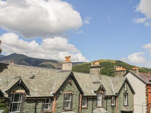 A row of traditional stone houses with pitched roofs and chimneys, set against a backdrop of green hills and a partly cloudy sky. - Sefton House (Keswick)