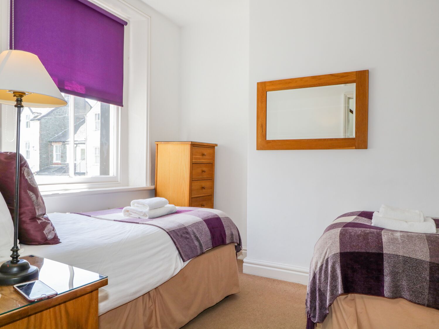 A hotel room with two beds, a wooden dresser, a mirror, and a window with a purple blind.