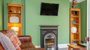 A living room with a green wall, a fireplace, a leather sofa, and a television mounted above the fireplace. - Sefton House (Keswick)
