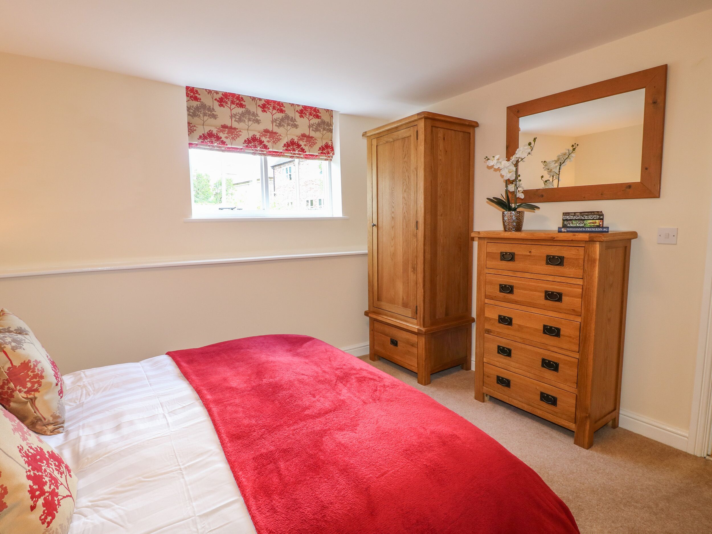 A bedroom with a bed, a wooden wardrobe, a dresser, a window with a floral blind, and a mirror on the wall.