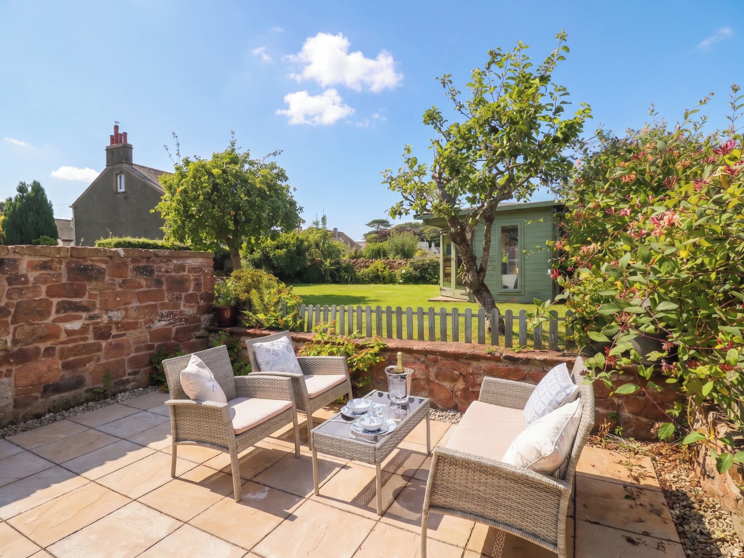 A patio with wicker furniture and a table, a stone wall, and a view of a green house and garden.