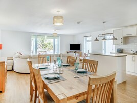 Uma cozinha moderna com armários brancos, uma sala de jantar com cadeiras de madeira e uma mesa posta para quatro pessoas, e uma TV na sala de estar.