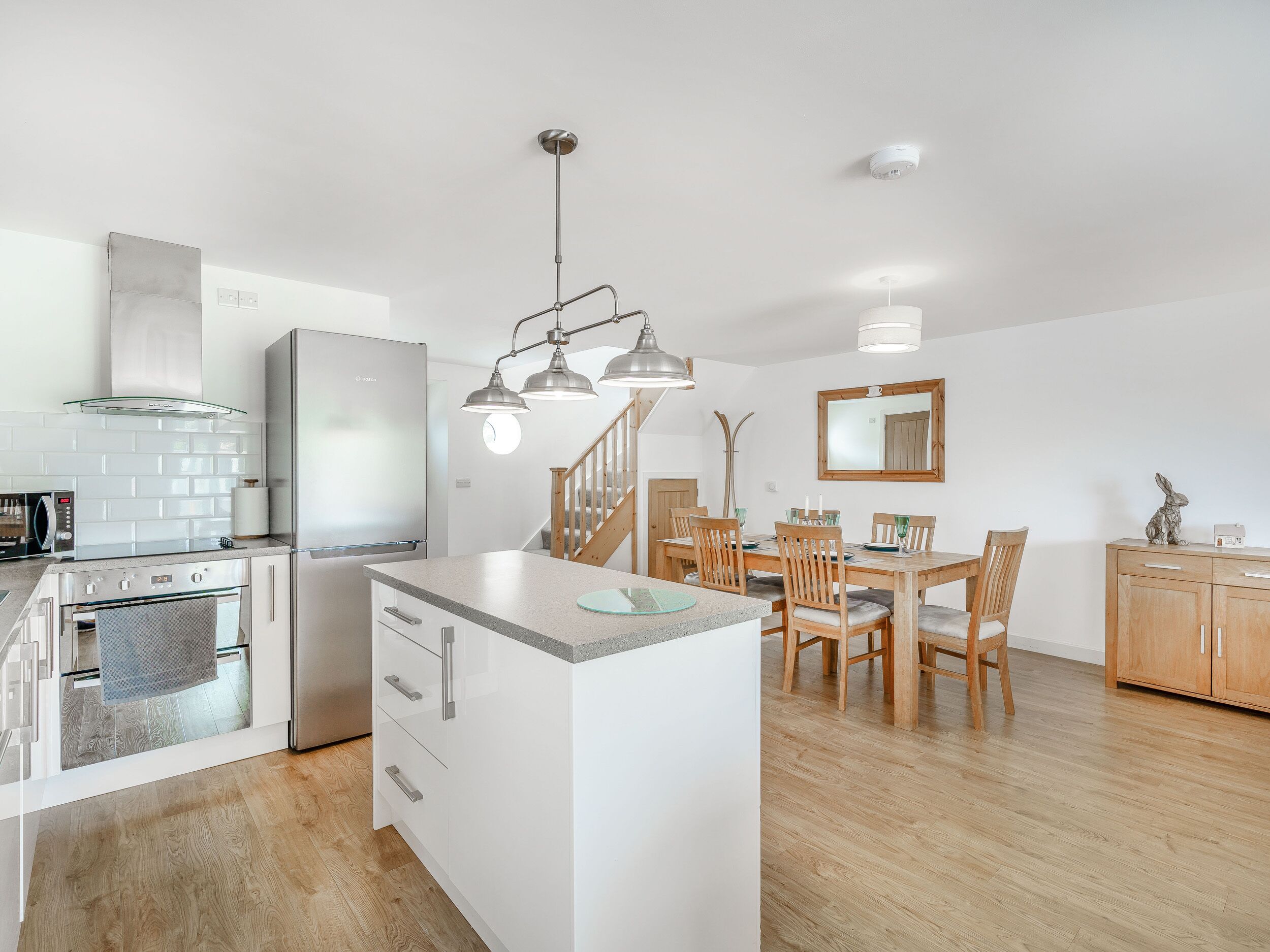 A modern kitchen with a central island, wooden dining table, and chairs. There is a built-in oven and a refrigerator.