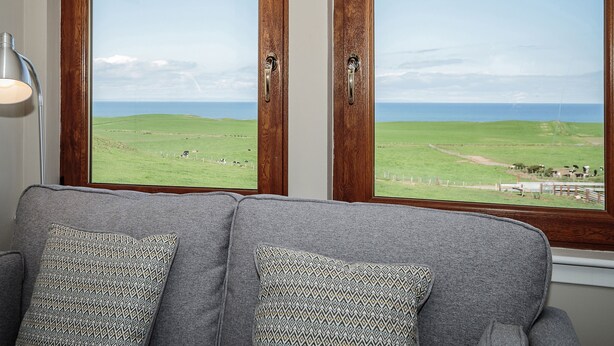 Ein Zimmer mit grauem Sofa, zwei gemusterten Kissen, einer Lampe und einem Fenster mit Blick auf ein grünes Feld und das Meer.