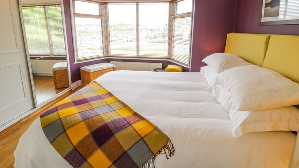Un dormitorio con una cama grande, una ventana con persianas, un pequeño banco y un cabecero amarillo.