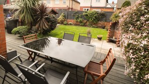 Un patio con una mesa y sillas de vidrio, rodeado por un jardín con diversas plantas y un muro de ladrillo.