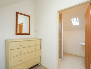 Ein Schlafzimmer mit einem beigen Schminktisch, einem Spiegel und einer Tür zu einem Badezimmer mit Waschbecken und Fenster.