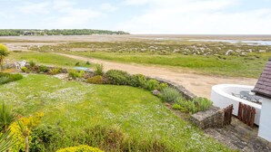 A coastal landscape with a grassy area, a white building with a curved roof, and a view of the sea and distant hills. - Pen y Lon (Pentraeth)