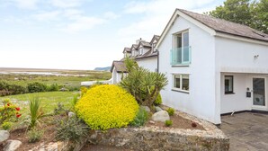 A modern two-story house with a white exterior, a stone retaining wall, and a well-maintained garden with yellow flowers and various shrubs. - Pen y Lon (Pentraeth)