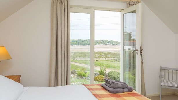 A bedroom with a bed, a window with curtains, a view of a landscape, and a small chair. - Pen y Lon (Pentraeth)