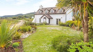 A white house with a sloped roof, a front porch, and a garden with various plants and a palm tree. - Pen y Lon (Pentraeth)