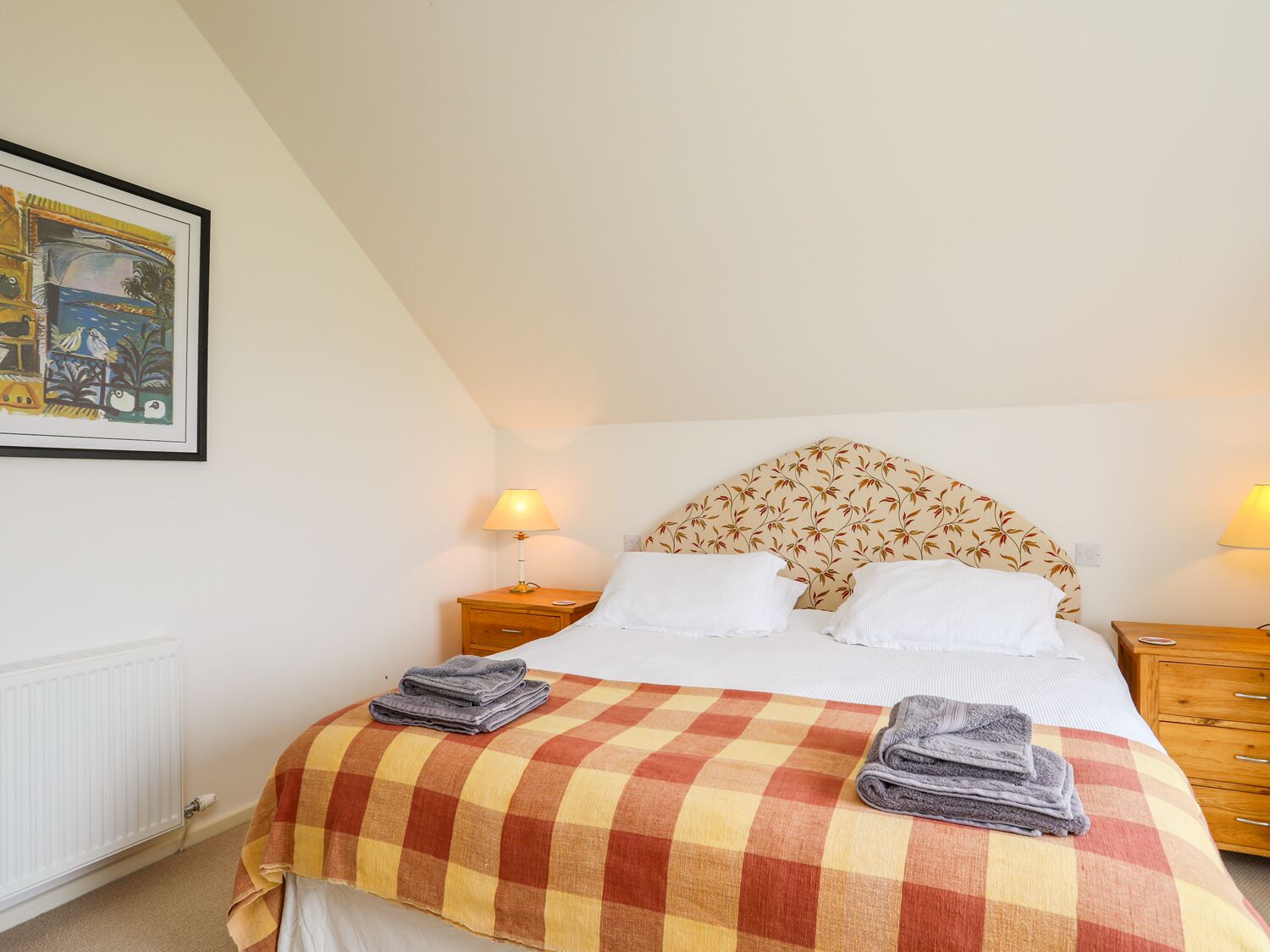A bedroom with a checkered bedspread, two bedside lamps, a headboard with a floral pattern, and a framed picture on the wall.