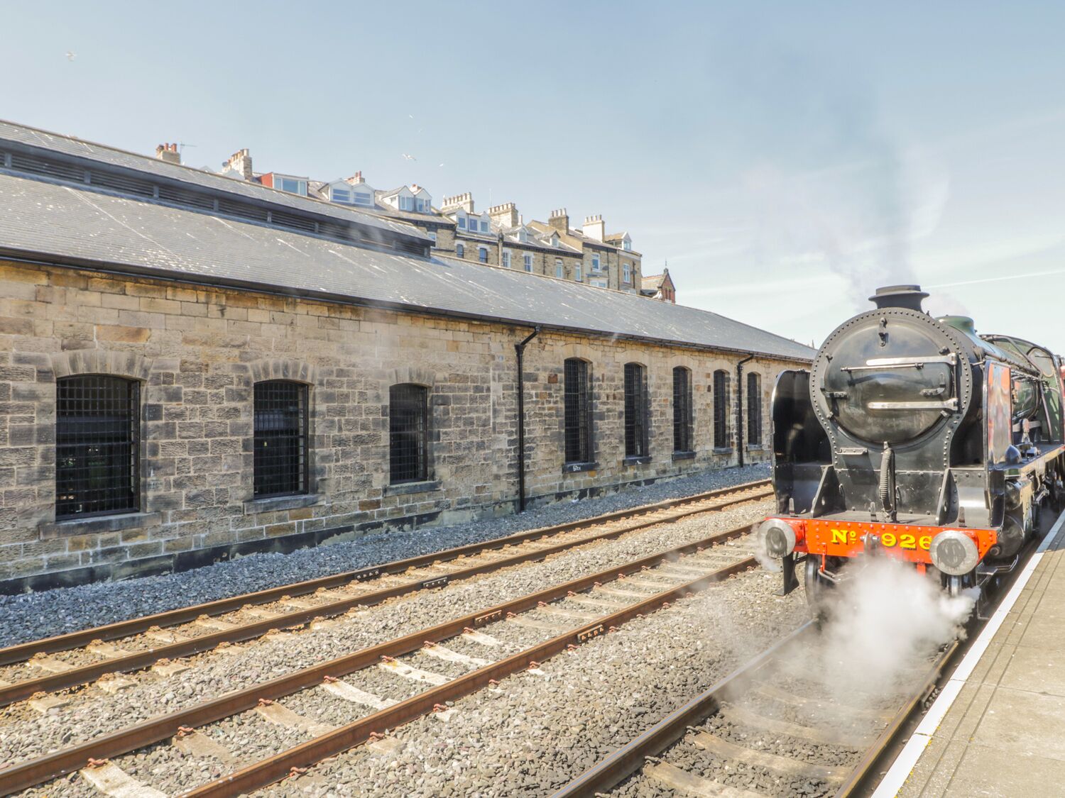 A steam locomotive at a railway station with stone buildings and multiple tracks.