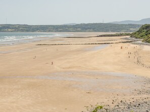 A wide sandy beach with a rocky shoreline, a few people scattered along the shore, and a distant view of hills and a tower. - Rhous (Tyn-y-Gongl)
