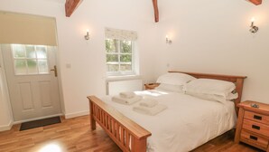 A bedroom with a wooden bed, white bedding, a wooden nightstand, a bedside table, a window with a view of greenery, and a white door. - Nursery Cottage (Morpeth)