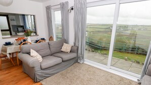 Ein gemütliches Wohnzimmer mit einer grauen Couch, einem Holzesstisch und Blick auf die Landschaft durch große Fenster.