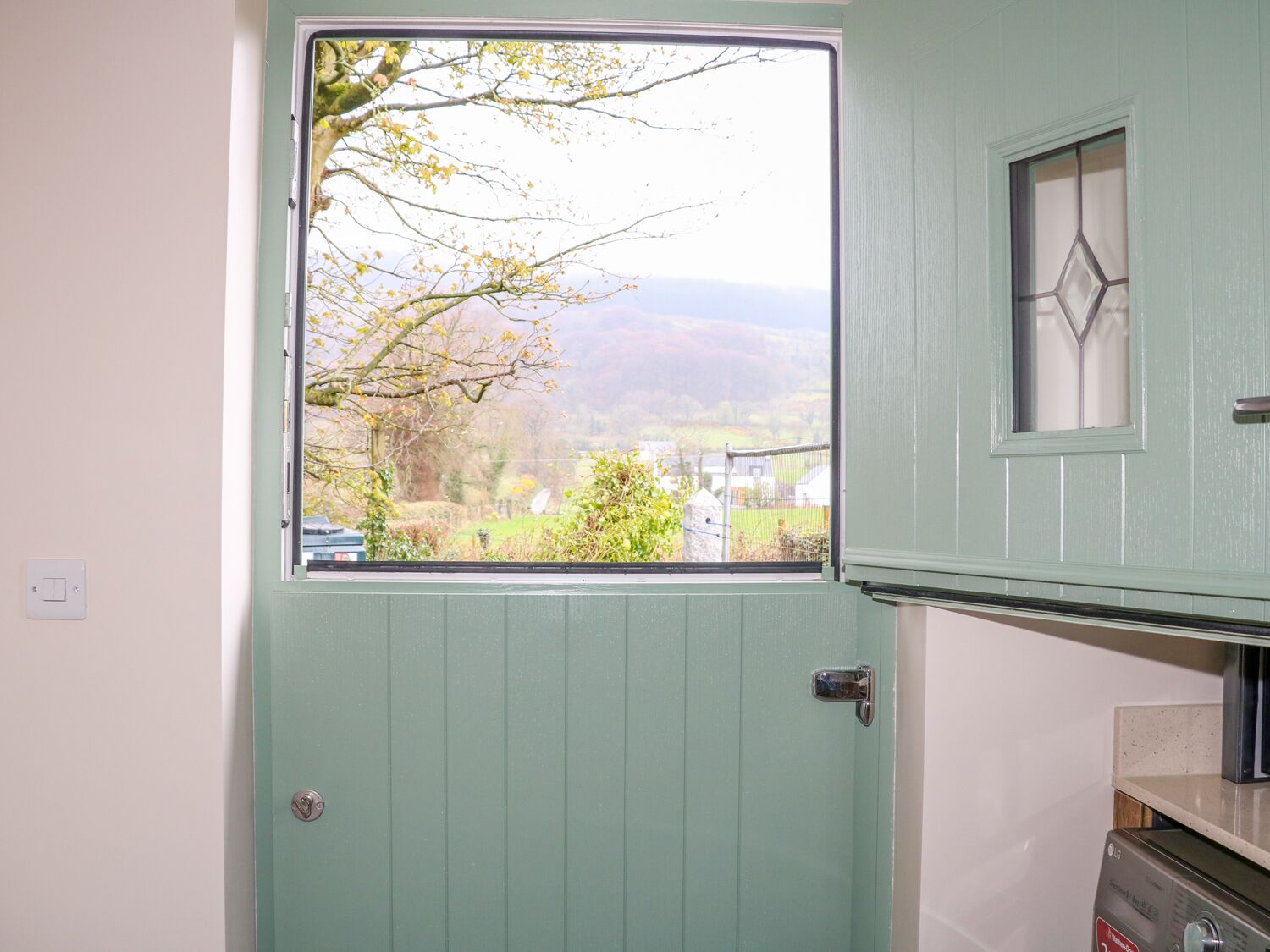 A room with a green door, a window with a view of a rural landscape, and a small cabinet with a mirror.