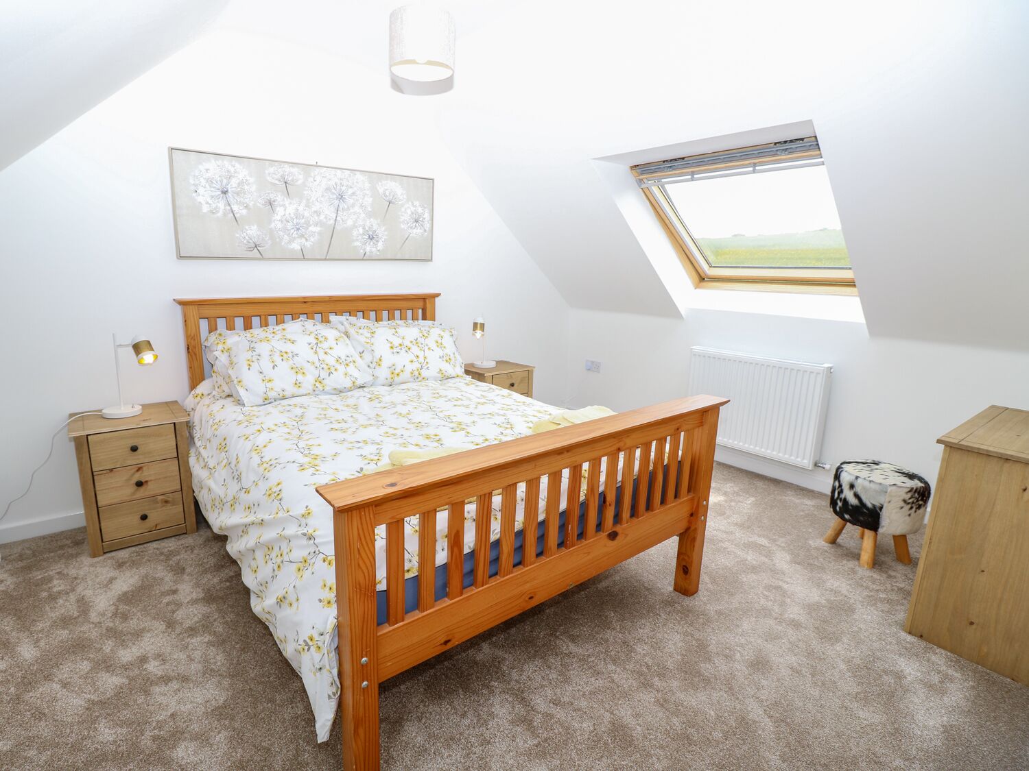 A bedroom with a wooden bed, a nightstand, a bedside lamp, a radiator, a small stool, and a skylight.
