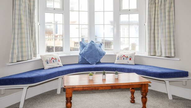 A room with a bay window seating area, white walls, and a wooden table. - The Pink House (Rhosneigr)